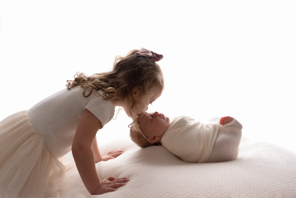 Backlit image of an older sister kissing forehead of newborn baby girl.