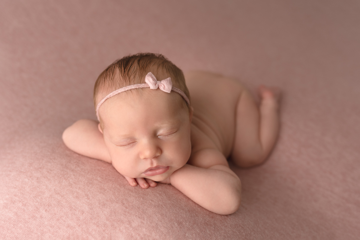 Baby girl laying on pink blanket on her tummy with her head on her hands, wearing a pink little bow headband.