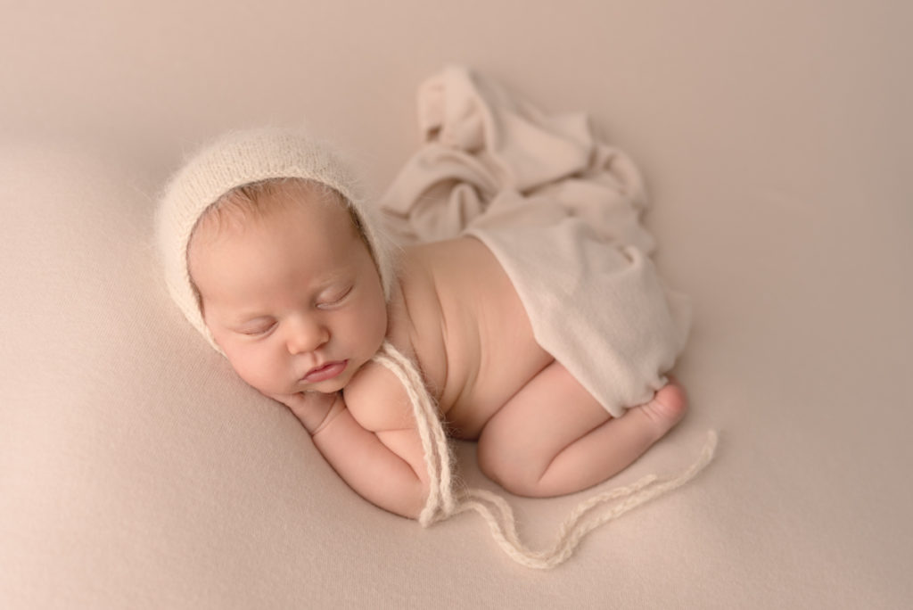 Newborn baby girl posed on her tummy with cream bonnet and cream wrap over bottom.