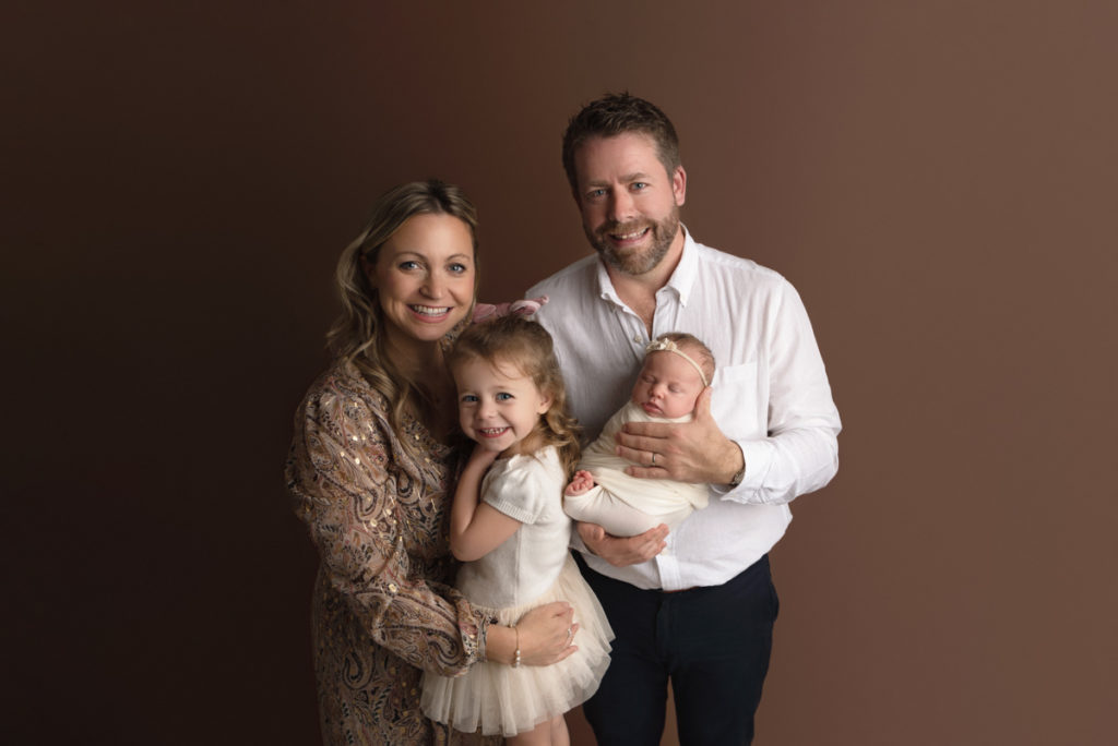 Family of 4 posing on a solid brown backdrop for a newborn family session