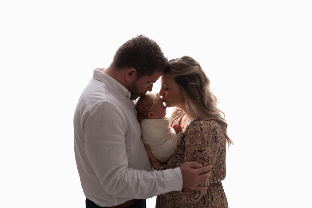 Backlit profile image of parents holding baby while both are kissing her forehead.