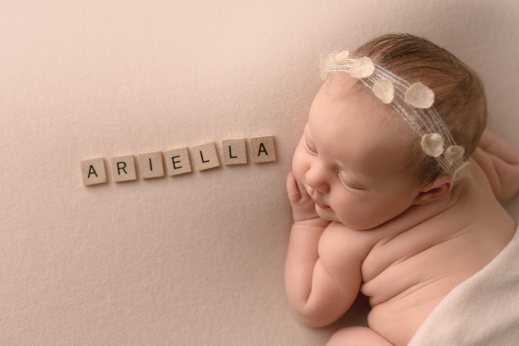 Newborn baby girl laying on tummy with name "Ariella" spelled out using scrabble tiles.