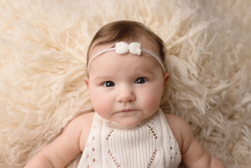 Close up of baby girl wearing white outfit and bow headband.  Laying on her back on a white flokati rug
