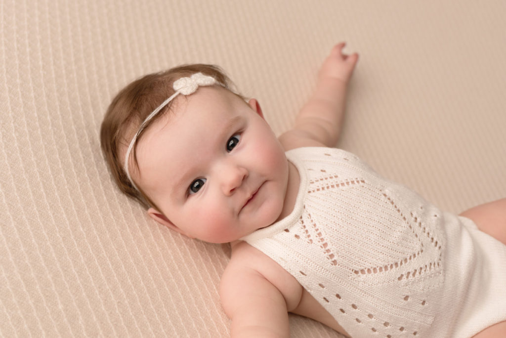 Close up image of baby girl in white outfit and bow, laying on a white blanket, smiling and looking at the camera
