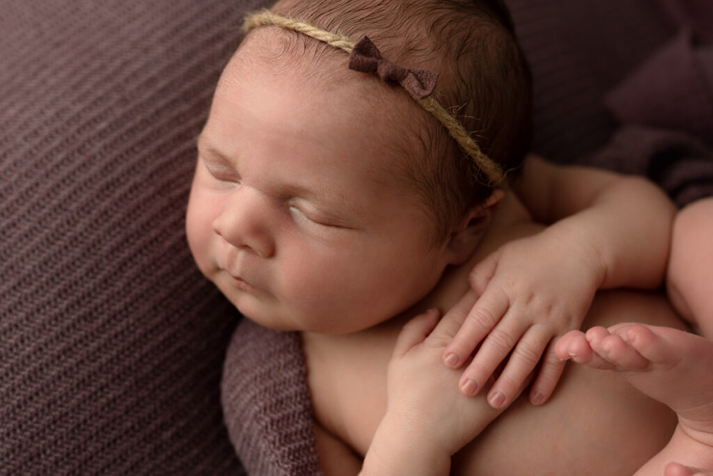 Close up profile image of newborn baby girl laying on her back on a dark purple blanket and purple little bow headband.
