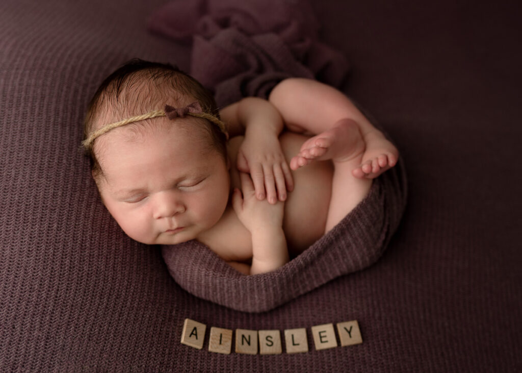 Newborn baby girl wrapped up in a dark purple wrap with scrabble letters spelling out Ainsley next to her.