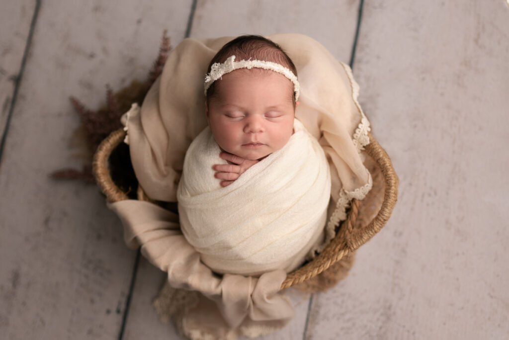 Newborn baby girl swaddled in white wrap, wearing a white bow headband and placed in a wicker basket.