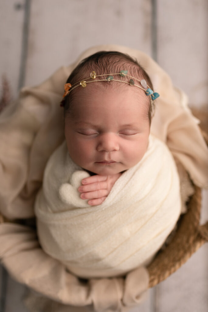 Close up image of newborn baby girl placed in a basket while wrapped in white swaddle, holding a white heart in hands and a rainbow jeweled headband.