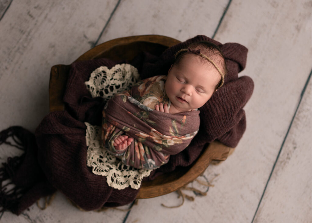 Newborn baby girl placed in a shallow wooden bowl, wrapped in a purple floral wrap with toes peeking out.