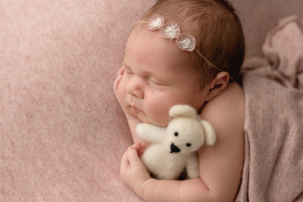 Close up of a newborn baby girl laying on her side, wrapped up in a pick swaddle and laying on a matching pink blanket.  She is holding a cream little teddy bear.