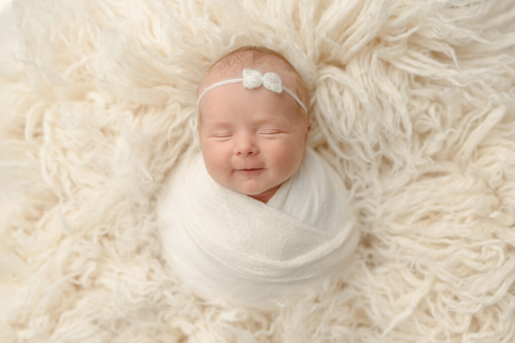 Newborn girl smiling swaddled in cream wrap laying on a cream flokati rug