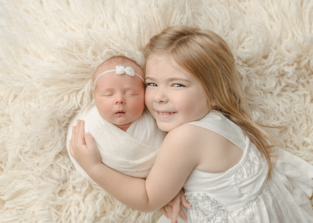 Newborn baby girl being cuddled by big sister, laying on a cream flokati rug