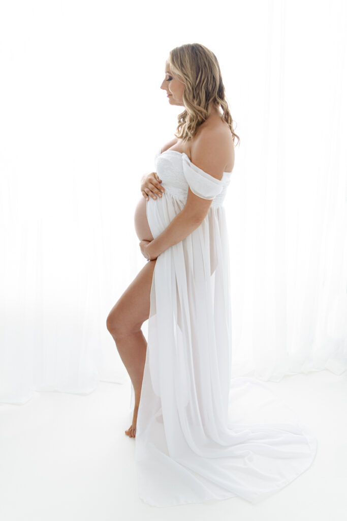 Backlit profile image of expecting mother in white lace gown, holding belly.
