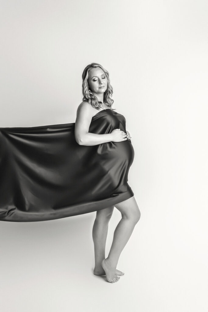 Black and white of expecting mom with black satin fabric draped across torso and legs.