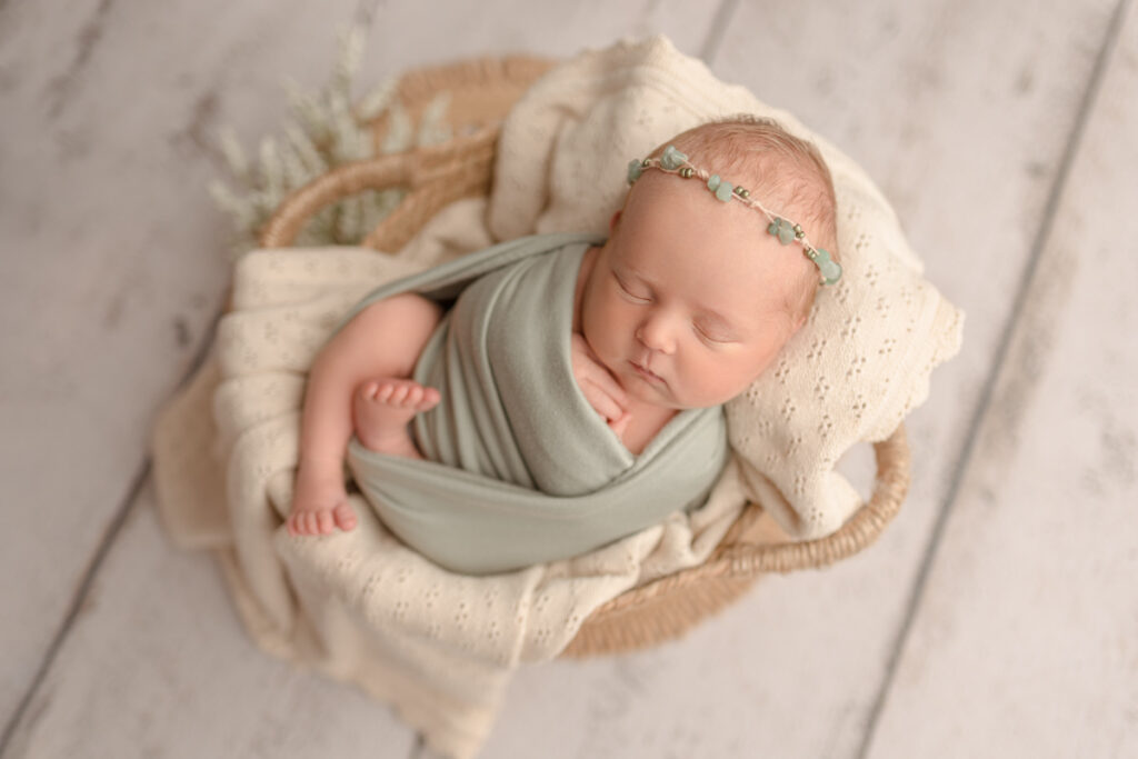 Newborn baby girl swaddled in a sage wrap, laying in a basket