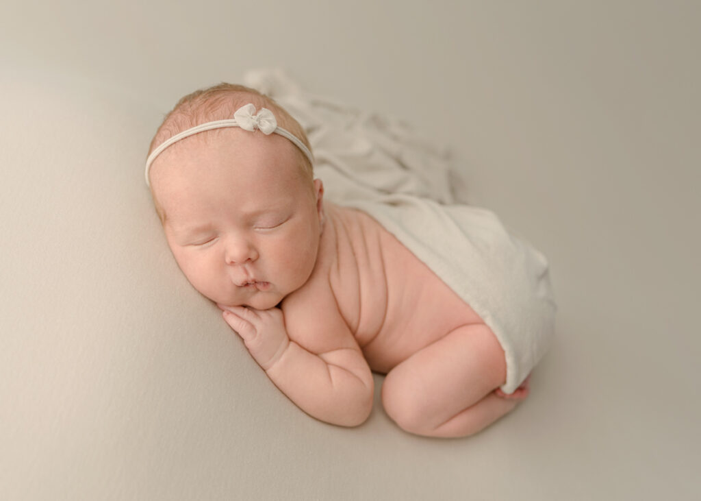 Newborn curled up laying tummy with tan swaddle, bow and blanket