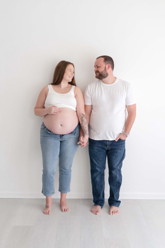 Expecting parents leaning on wall holding hands while looking towards each other.