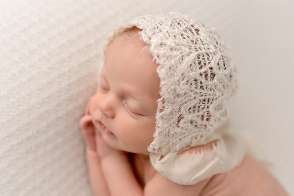 Close up image of newborn baby girl sleeping on her side with her hands under her cheeks, wearing a lace white bonnet.