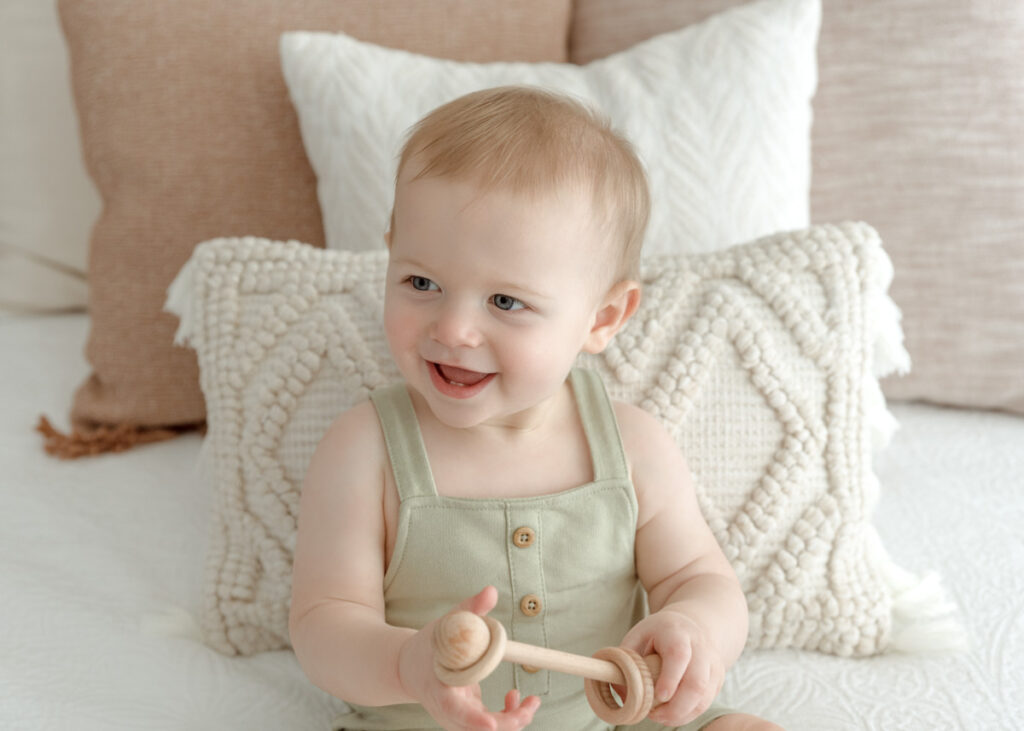Close up image of milestone boy sitting on a bed smiling, holding a rattle, wearing green overalls.