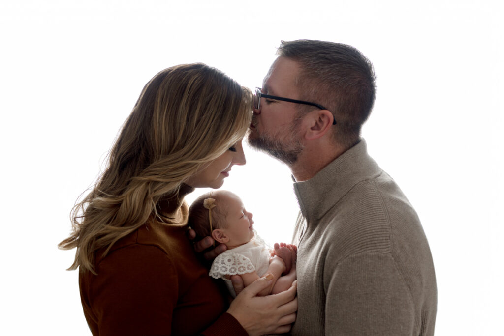 Backlit profile image capturing parents holding newborn baby girl, while dad is kissing mom's forehead.