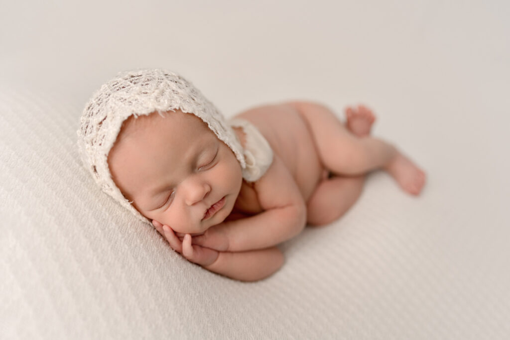 Newborn baby girl laying on her side wearing a lace white bonnet with her hands under her cheek.