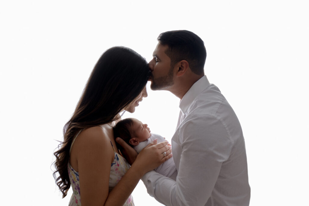 Backlit profile image of new parents holding their baby boy while dad kisses mom's forehead.