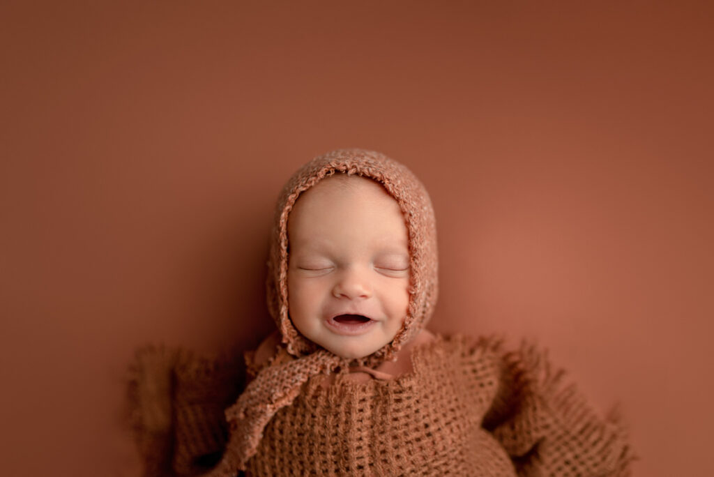 Newborn baby girl wrapped up in a rust wrap and bonnet, laying on her back and smiling at the camera.