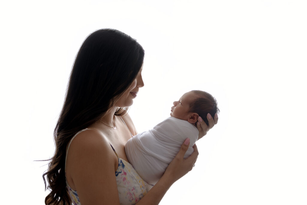 Backlit profile image of new. mom holding out her baby boy while looking down at him.