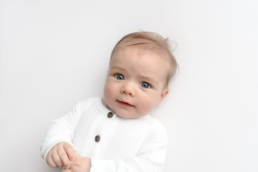 4 month old baby dressed in white button up outfit, laying on a white backdrop, smiling at camera.

