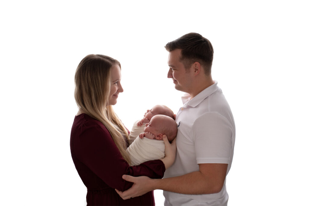 Gulf Breeze, FL newborn photographer.  Backlit profile image of new parents holding their twin babies and smiling at each other during a posed studio photography session.