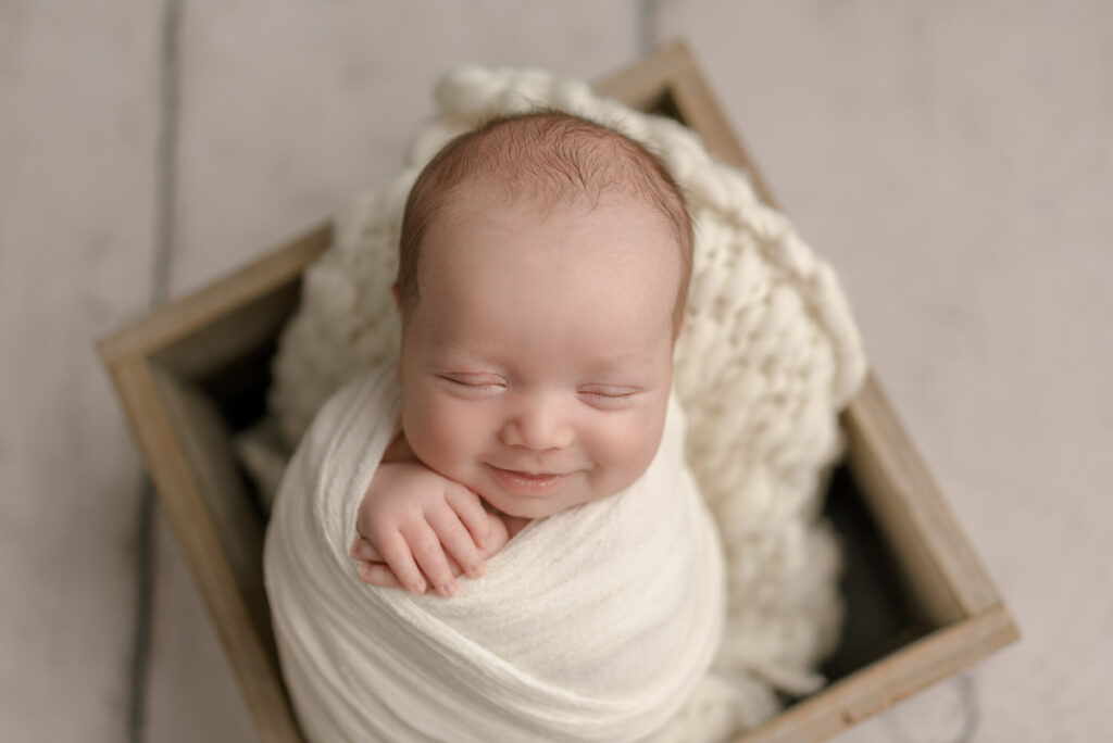 Milton, FL newborn photographer. Baby sleeping while swaddled in cream placed in wooden box prop and smiling during a studio posed photography session.
