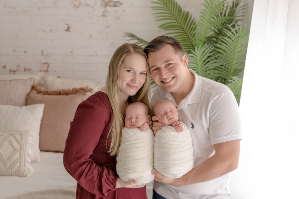 Pensacola, FL newborn photographer.  New parents posing and holding their baby boy twins looking at the camera smiling during a lifestyle studio photography session.