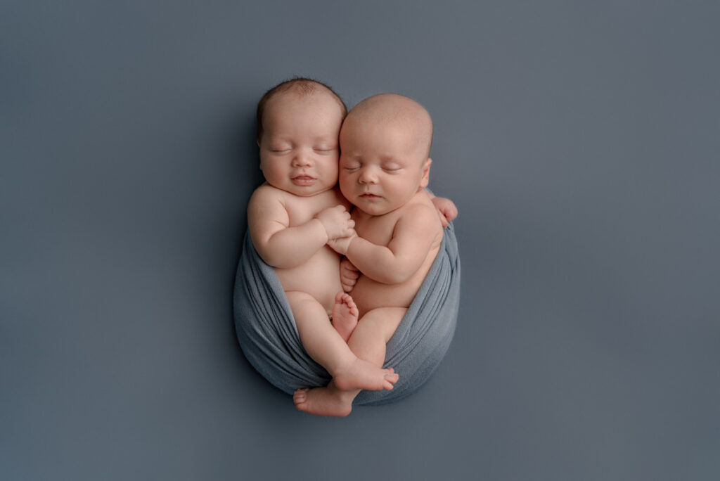 Pensacola, FL newborn photographer.  Baby boy twins swaddled together, snuggling while wrapped in a blue wrap laying down on a blue backdrop during a posed studio photography session.